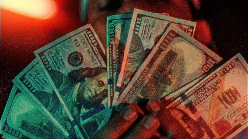 A person holds up several spread-out $100 bills, fanned in front of their face, with a warm red glow in the background—illustrating potential benefits from the HITS Act.