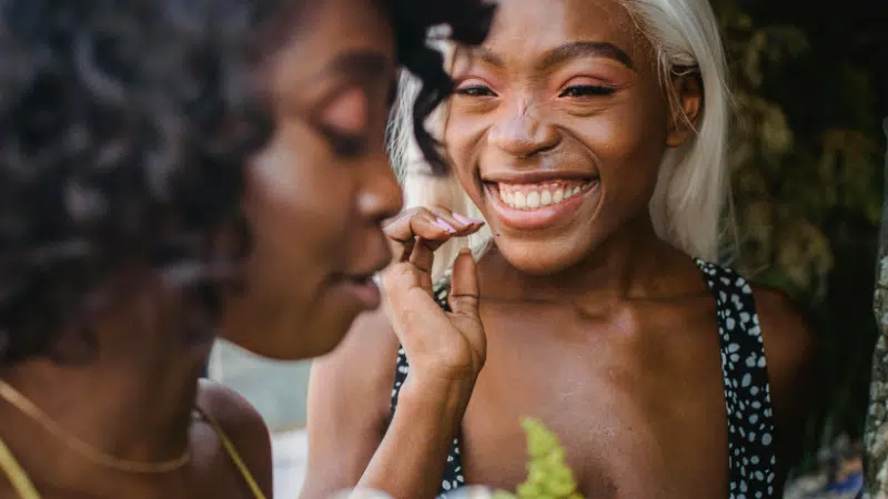Two women are outdoors; one is smiling broadly at the camera while the other stands nearby, facing her. Both appear relaxed and happy, enjoying a moment that reflects the positive impact of nurturing mental health.