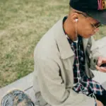 A person sitting on a concrete ledge outdoors, wearing a cap, glasses, headphones, and a beige jacket, looking at a smartphone—perhaps checking Spotify. Grass is visible in the background.