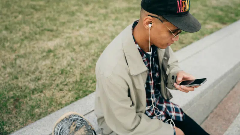 A person sitting on a concrete ledge outdoors, wearing a cap, glasses, headphones, and a beige jacket, looking at a smartphone—perhaps checking Spotify. Grass is visible in the background.