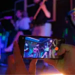 A person holds a smartphone, recording or photographing a live band performing on stage under colorful lights—perfect content to share on YouTube for Artists.