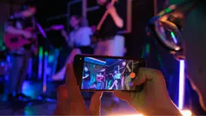 A person holds a smartphone, recording or photographing a live band performing on stage under colorful lights—perfect content to share on YouTube for Artists.
