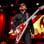 A musician wearing dark clothes and sunglasses plays a red electric guitar and sings on stage, with a dragon statue displayed on a screen behind him at the Latin GRAMMY awards.