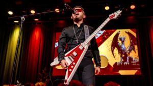 A musician wearing dark clothes and sunglasses plays a red electric guitar and sings on stage, with a dragon statue displayed on a screen behind him at the Latin GRAMMY awards.