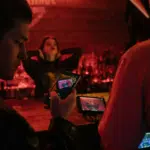 Two people film a woman posing at a dimly lit bar, ready to capture her best look before release, using a smartphone and a camera with a monitor.