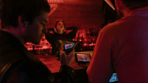 Two people film a woman posing at a dimly lit bar, ready to capture her best look before release, using a smartphone and a camera with a monitor.