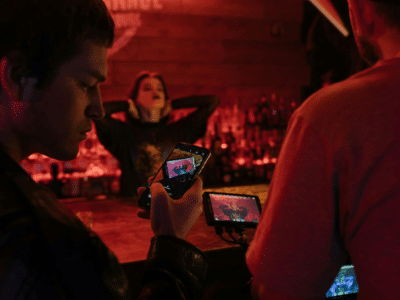 Two people film a woman posing at a dimly lit bar, ready to capture her best look before release, using a smartphone and a camera with a monitor.