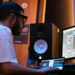 A person wearing glasses sits at a desk, working on music production software for their label on a computer in a studio with speakers and dim lighting.