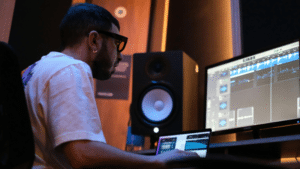 How to Build Your Brand as an Independent Label A person wearing glasses sits at a desk, working on music production software for their label on a computer in a studio with speakers and dim lighting.
