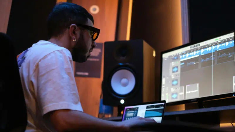 A person wearing glasses sits at a desk, working on music production software for their label on a computer in a studio with speakers and dim lighting.