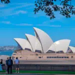People stand and walk near the Sydney Opera House in Australia, with its distinctive white sail-shaped roofs, under a clear blue sky.
