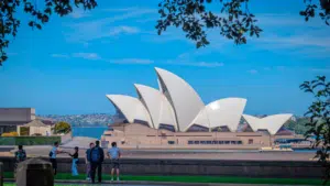 People stand and walk near the Sydney Opera House in Australia, with its distinctive white sail-shaped roofs, under a clear blue sky.