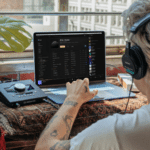 Person with headphones using a laptop at a desk near a window, interacting with audio equipment and music software.