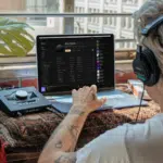 Person with headphones using a laptop at a desk near a window, interacting with audio equipment and music software.