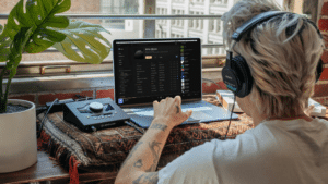 SEO for Musicians: Why It Matters and How To Use It Person with headphones using a laptop at a desk near a window, interacting with audio equipment and music software.