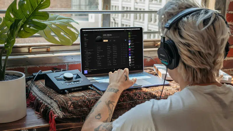 Person with headphones using a laptop at a desk near a window, interacting with audio equipment and music software.