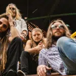 Four men with long hair and sunglasses sit on steps outdoors, looking at the camera—perfect for a playlist cover. The background features yellow and green railings and a wooden structure.
