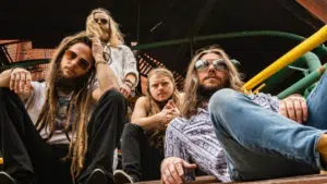 Four men with long hair and sunglasses sit on steps outdoors, looking at the camera—perfect for a playlist cover. The background features yellow and green railings and a wooden structure.