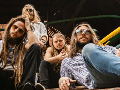 Four men with long hair and sunglasses sit on steps outdoors, looking at the camera—perfect for a playlist cover. The background features yellow and green railings and a wooden structure.