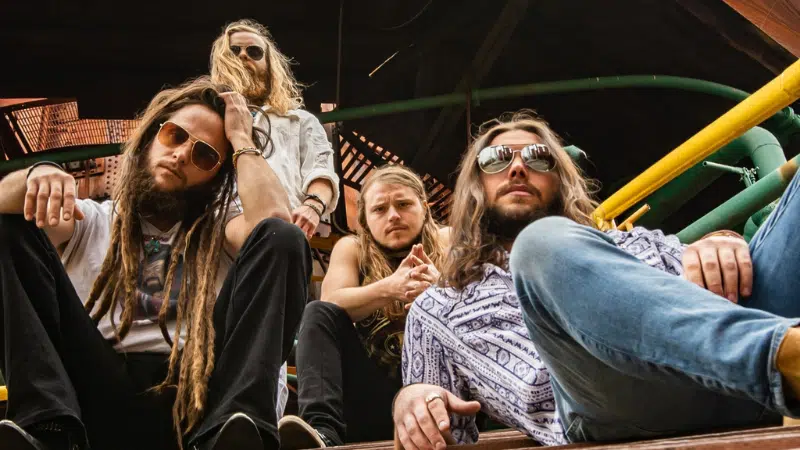 Four men with long hair and sunglasses sit on steps outdoors, looking at the camera—perfect for a playlist cover. The background features yellow and green railings and a wooden structure.