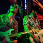 Two guitarists play electric guitars on stage under colorful lights at festivals, both wearing patterned shirts and wristbands, with a vivid background behind them.