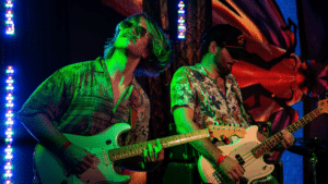 Two guitarists play electric guitars on stage under colorful lights at festivals, both wearing patterned shirts and wristbands, with a vivid background behind them.