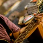 In San Juan, a person wearing a hat and glasses plays an acoustic guitar, focusing on forming chords with their left hand; music equipment is visible in the background.