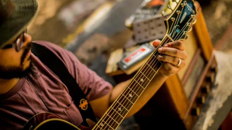 In San Juan, a person wearing a hat and glasses plays an acoustic guitar, focusing on forming chords with their left hand; music equipment is visible in the background.