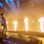 A DJ performs on stage with flames shooting up in front of a large, energetic crowd of fans at a concert.