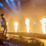A DJ performs on stage with flames shooting up in front of a large, energetic crowd of fans at a concert.
