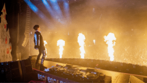 A DJ performs on stage with flames shooting up in front of a large, energetic crowd of fans at a concert.