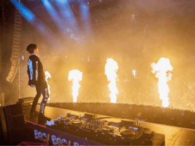A DJ performs on stage with flames shooting up in front of a large, energetic crowd of fans at a concert.
