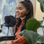 A woman with long dark hair sits by a microphone, smiling and speaking, surrounded by green plants in a well-lit indoor space, exuding PR expertise.