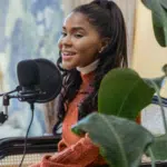 A woman with long dark hair sits by a microphone, smiling and speaking, surrounded by green plants in a well-lit indoor space, exuding PR expertise.