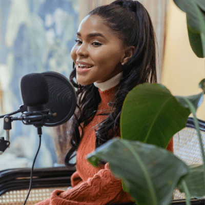 A woman with long dark hair sits by a microphone, smiling and speaking, surrounded by green plants in a well-lit indoor space, exuding PR expertise.