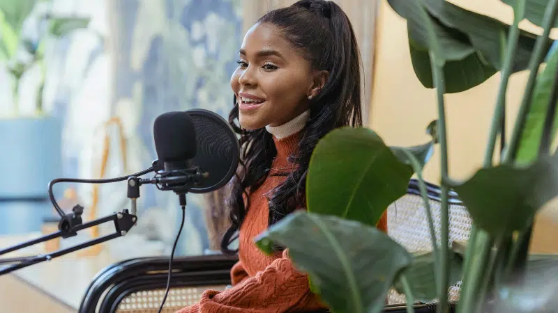 A woman with long dark hair sits by a microphone, smiling and speaking, surrounded by green plants in a well-lit indoor space, exuding PR expertise.