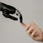 A robotic hand and a human hand reach toward each other, with their fingers nearly touching against a plain background, symbolizing the intersection of technology and humanity in a scene reminiscent of iconic copyright imagery.