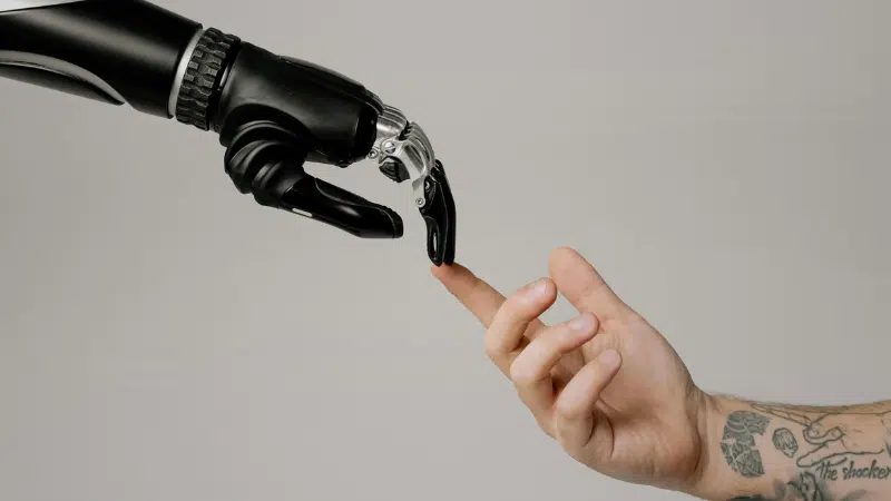 A robotic hand and a human hand reach toward each other, with their fingers nearly touching against a plain background, symbolizing the intersection of technology and humanity in a scene reminiscent of iconic copyright imagery.