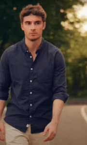 A man in a navy button-up shirt and light pants walks outdoors on a curved path with trees and songtools in the background.