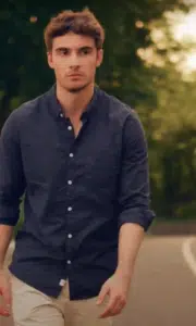 A man in a navy button-up shirt and light pants walks outdoors on a curved path with trees and songtools in the background.
