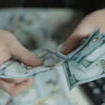 A person counts several US $100 bills over a desk with a calculator in the background, possibly preparing to handle taxes.