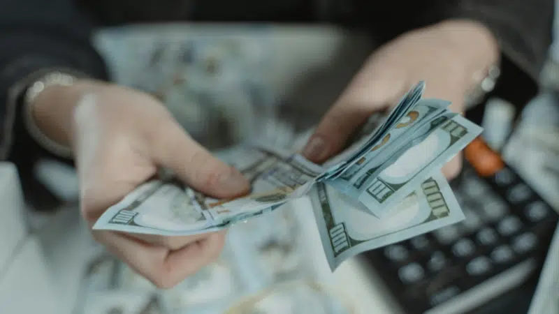 A person counts several US $100 bills over a desk with a calculator in the background, possibly preparing to handle taxes.