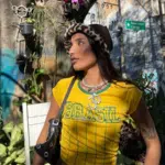 A woman wearing a yellow "Brasil" shirt, a leopard print hat, and a black shoulder bag stands outdoors among plants, with a sunlit wall and street sign in the background.