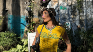 A woman wearing a yellow "Brasil" shirt, a leopard print hat, and a black shoulder bag stands outdoors among plants, with a sunlit wall and street sign in the background.