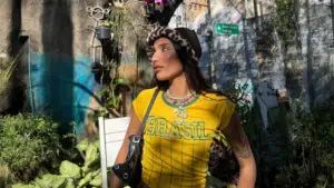 A woman wearing a yellow "Brasil" shirt, a leopard print hat, and a black shoulder bag stands outdoors among plants, with a sunlit wall and street sign in the background.