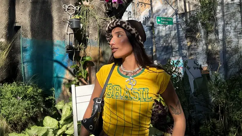 A woman wearing a yellow "Brasil" shirt, a leopard print hat, and a black shoulder bag stands outdoors among plants, with a sunlit wall and street sign in the background.