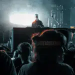 A DJ performing on stage with electronic equipment, illuminated by bright lights, as an audience watches in the foreground—music filling the air like a kind of therapy at a vibrant concert or music event.