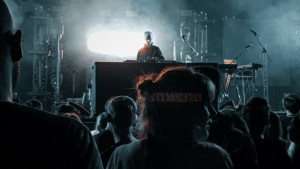 A DJ performing on stage with electronic equipment, illuminated by bright lights, as an audience watches in the foreground—music filling the air like a kind of therapy at a vibrant concert or music event.