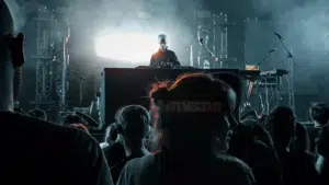 A DJ performing on stage with electronic equipment, illuminated by bright lights, as an audience watches in the foreground—music filling the air like a kind of therapy at a vibrant concert or music event.