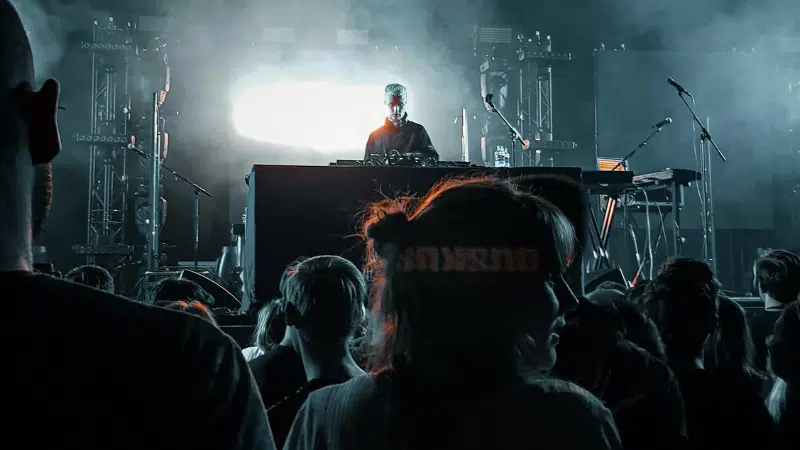 A DJ performing on stage with electronic equipment, illuminated by bright lights, as an audience watches in the foreground—music filling the air like a kind of therapy at a vibrant concert or music event.